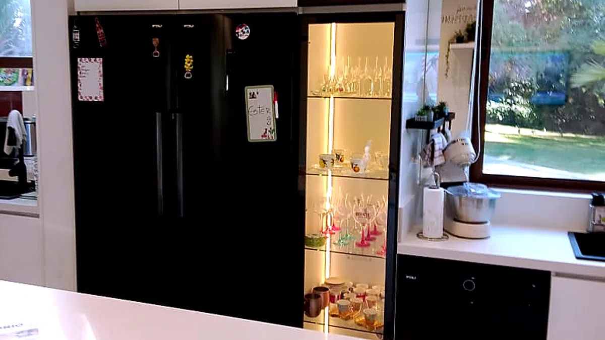 Cocina terminada con refrigerador negro, una vitrina iluminada con cristalería y mesón blanco despejado junto a una ventana con vista al jardín.