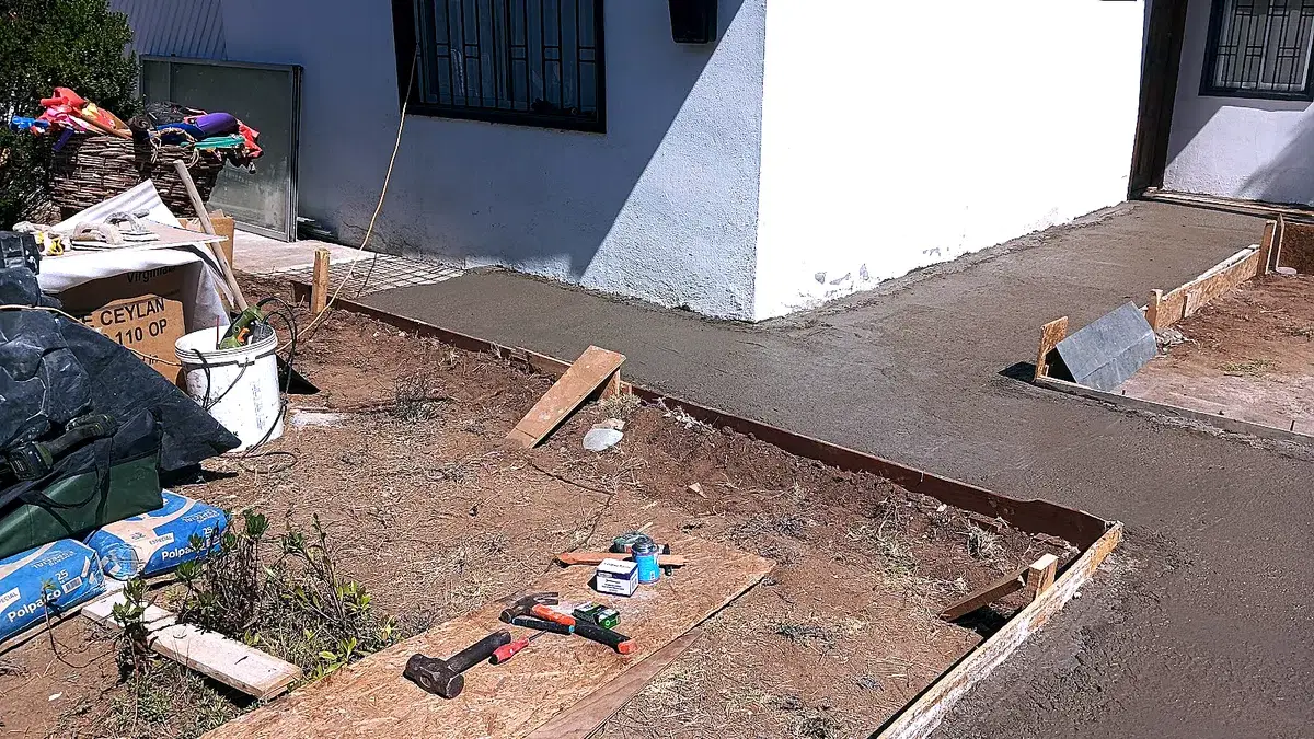 Radier de hormigón recién vaciado junto a la fachada de una casa, con el moldaje de madera aún puesto, sacos de cemento y herramientas (martillo, prensa y nivel) apoyadas en el suelo.