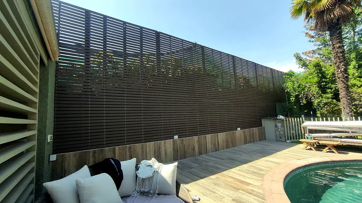 Terraza terminada con deck de porcelanato tipo madera, una celosía de listones de madera oscura como cierre, un living exterior con cojines y una piscina a un costado.
