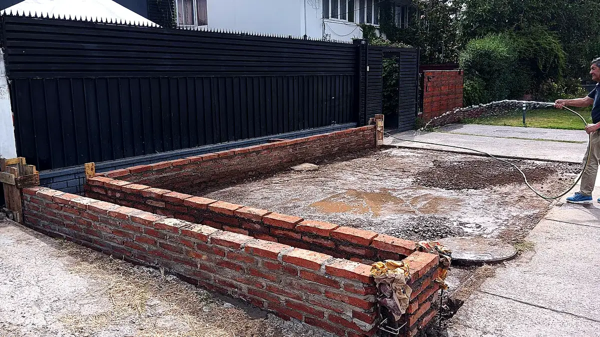 Trabajo de radier y pavimento en el acceso de una vivienda, con muro perimetral de ladrillo, portón metálico y un operario mojando la superficie con manguera.