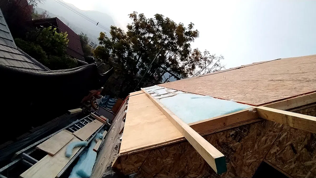Vista desde abajo de la techumbre de una casa en construcción: tableros estructurales de madera fijados sobre las vigas del techo, con aislante y material de obra apilado a un costado.