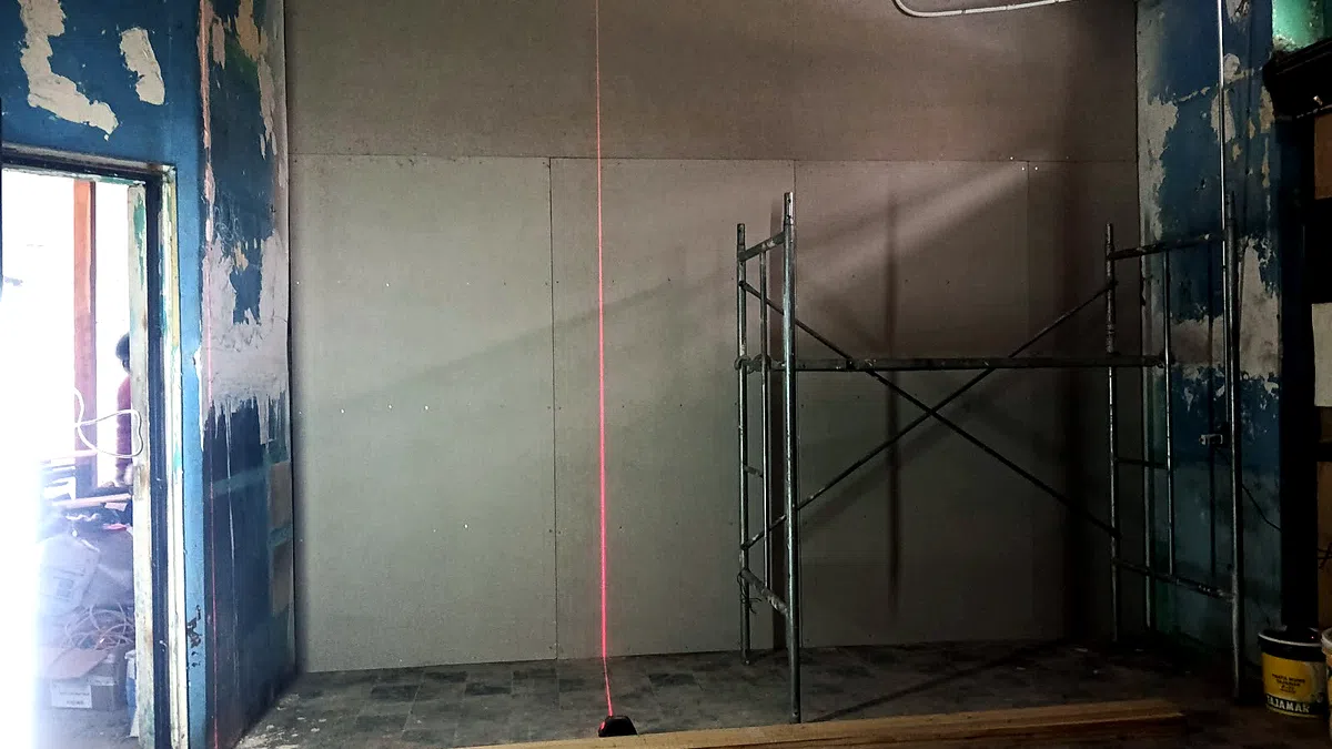 Interior de una vivienda en remodelación: un tabique nuevo de tablero ya montado y aplomado con nivel láser, andamios de trabajo y los muros antiguos con la pintura descascarada alrededor.